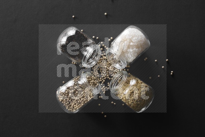 4 glass spice jars full of salt, black peppers, white peppers and cumin on black flooring