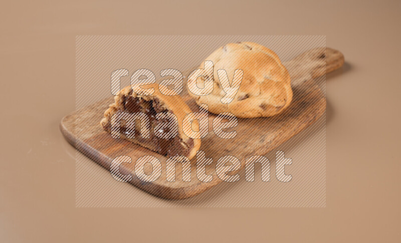 a chocolate chip cookie with another one cut in half on a wooden cutting board on a brown background