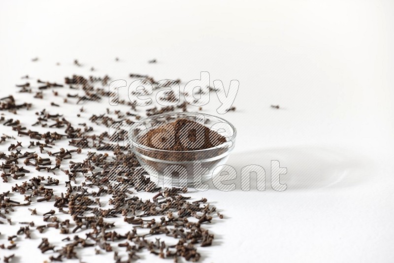 A glass bowl full of cloves powder and cloves grains spread on white flooring