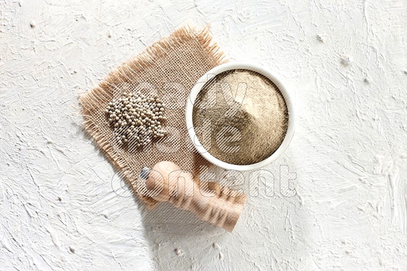 White pottery bowl full of white pepper powder set on a burlap piece of fabric with pepper beads and wooden pepper grinder on textured white flooring