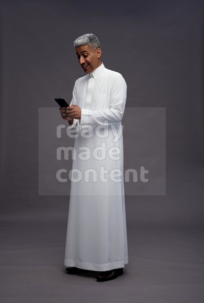 Saudi man wearing thob standing texting on phone on gray background
