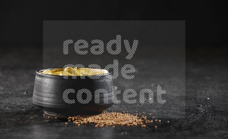 A black pottery bowl full of mustard paste with mustard seeds underneath on textured black flooring