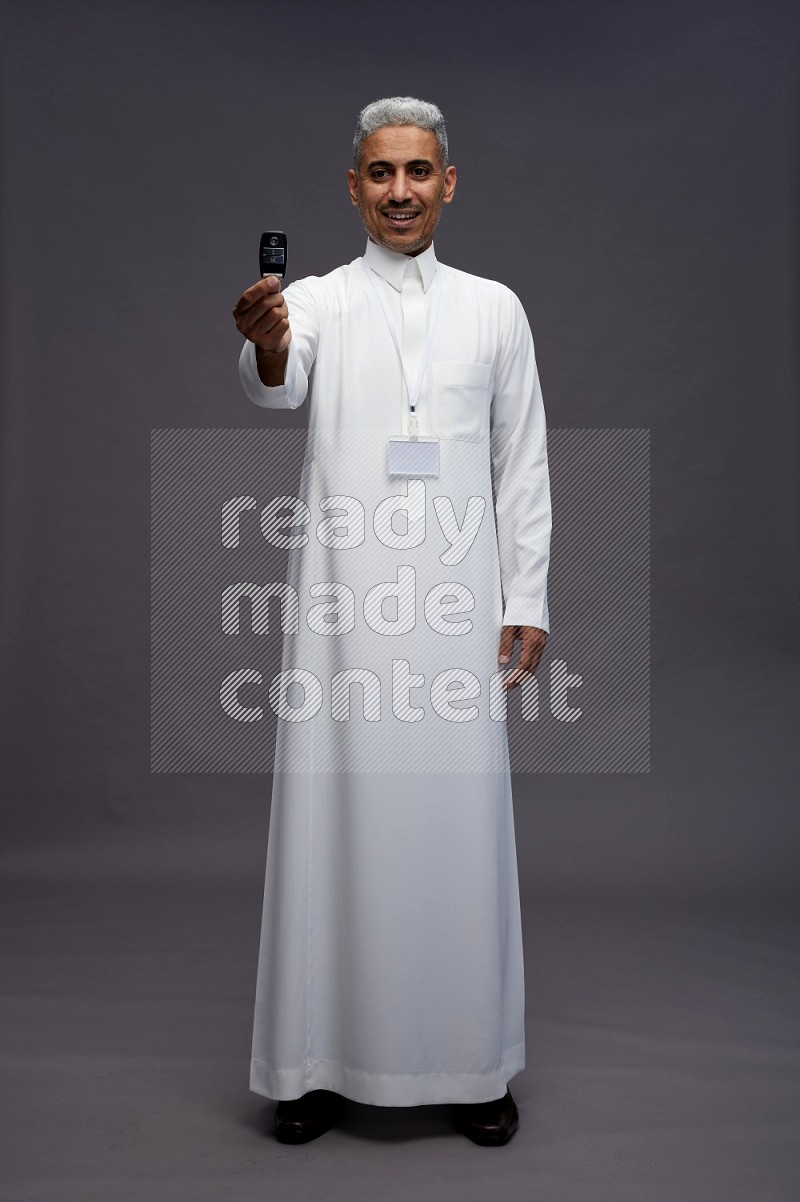 Saudi man wearing thob with neck strap employee badge standing holding key car on gray background