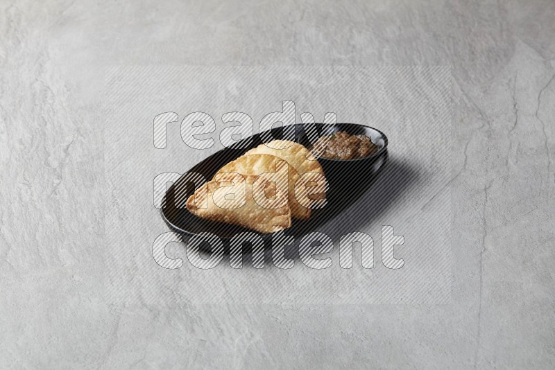Three fried sambosas in an oval shaped black plate and a sauce in a black round ramekin on a gray background
