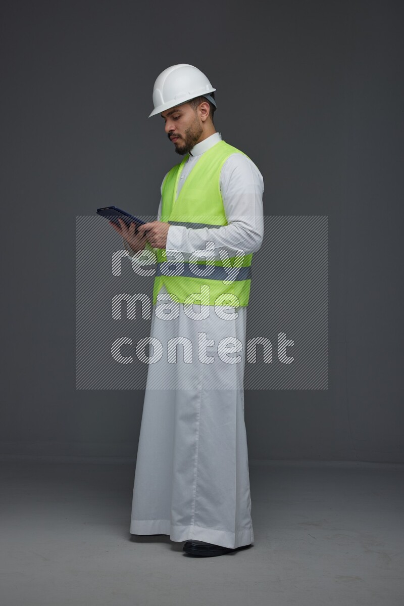 A Saudi man wearing Thobe with a yellow safety vest and white helmet standing and using his tablet different angles eye level on a gray background