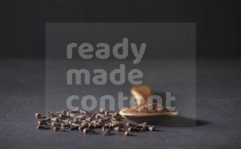A wooden ladle full of cloves powder and some of whole cloves around it on a black flooring