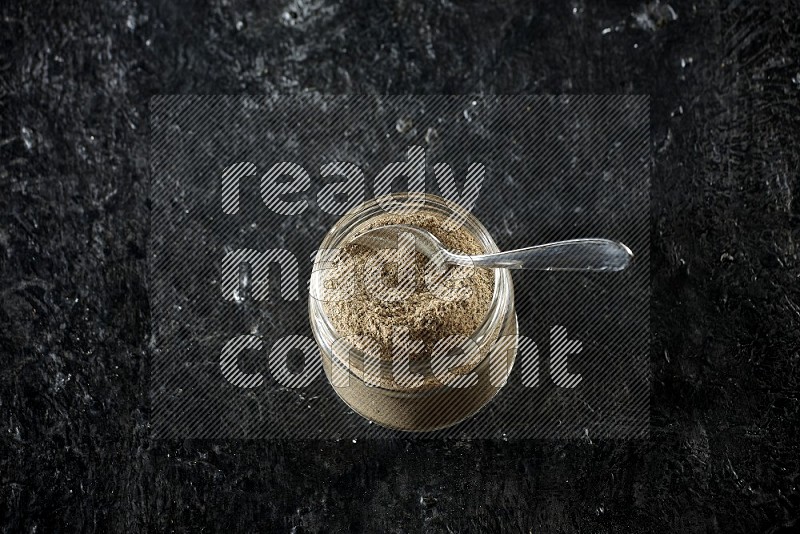 A glass jar and metal spoon full of cardamom powder on textured black flooring