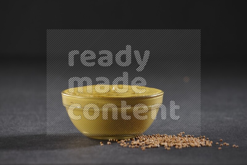 A glass bowl full of mustard paste with mustard seeds underneath on black flooring