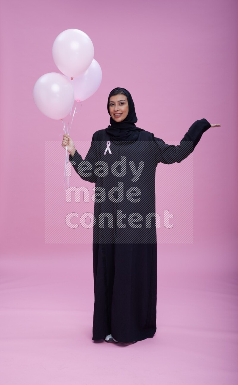 Saudi woman wearing pink ribbon on Abaya standing holding balloons on plain pink background