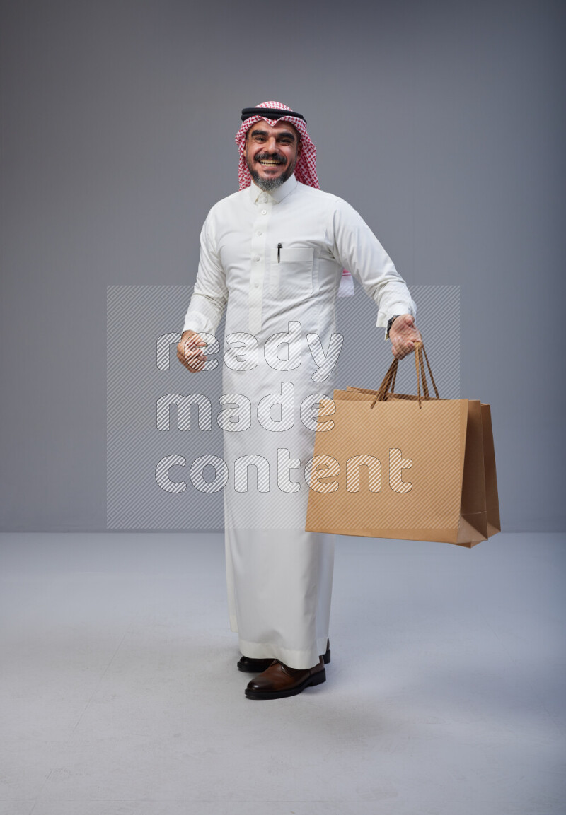 Saudi man Wearing Thob and red Shomag standing holding shopping bag on Gray background