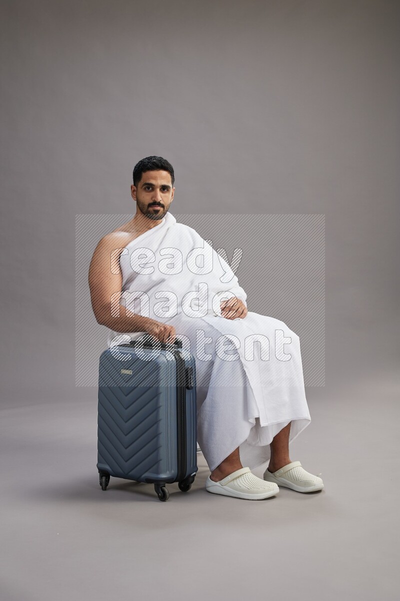 A man wearing Ehram sitting on chair holding traveling bag on gray background