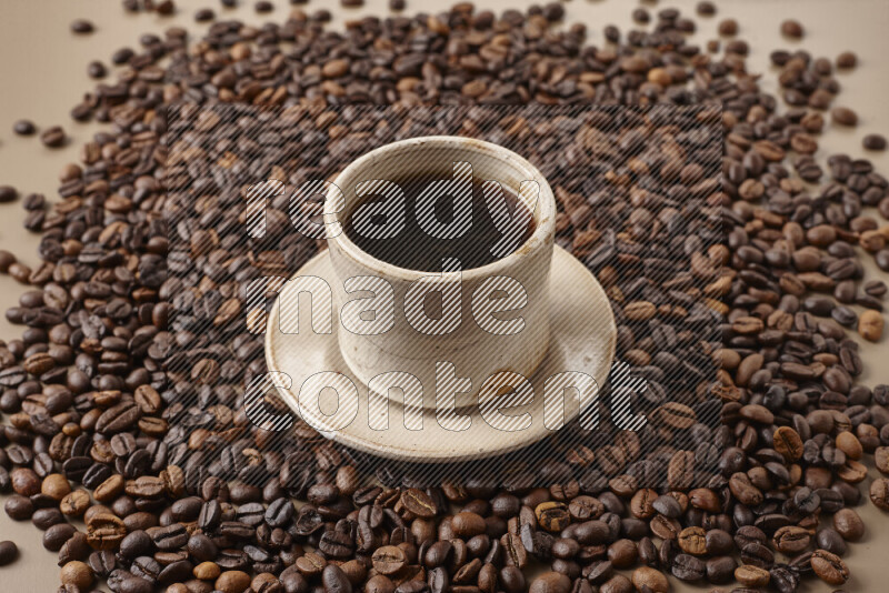 A beige pottery cup of coffee surrounded by roasted coffee beans on beige background