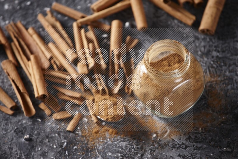 Herbal glass jar and a metal spoon full of cinnamon powder surrounded by cinnamon sticks on textured black background