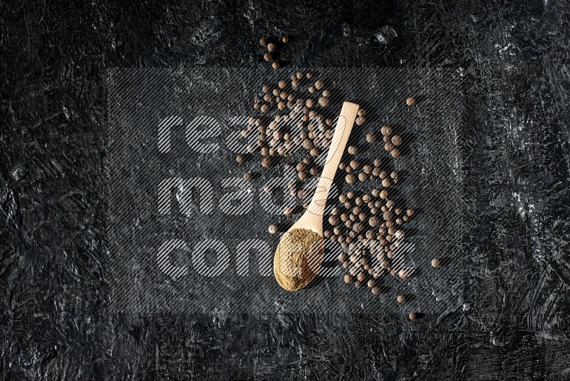 Wooden spoons full of all spice powder and allspice whole balls beside it on a textured black flooring