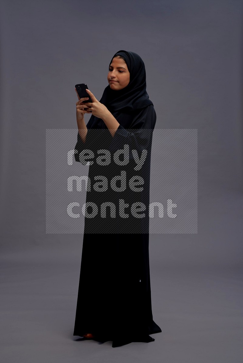 Saudi woman wearing Abaya standing texting on phone on gray background