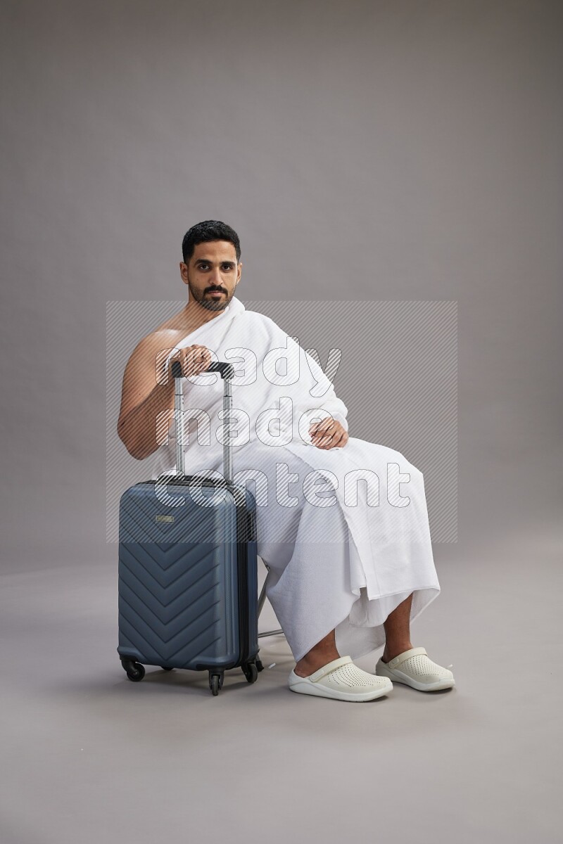 A man wearing Ehram sitting on chair holding traveling bag on gray background