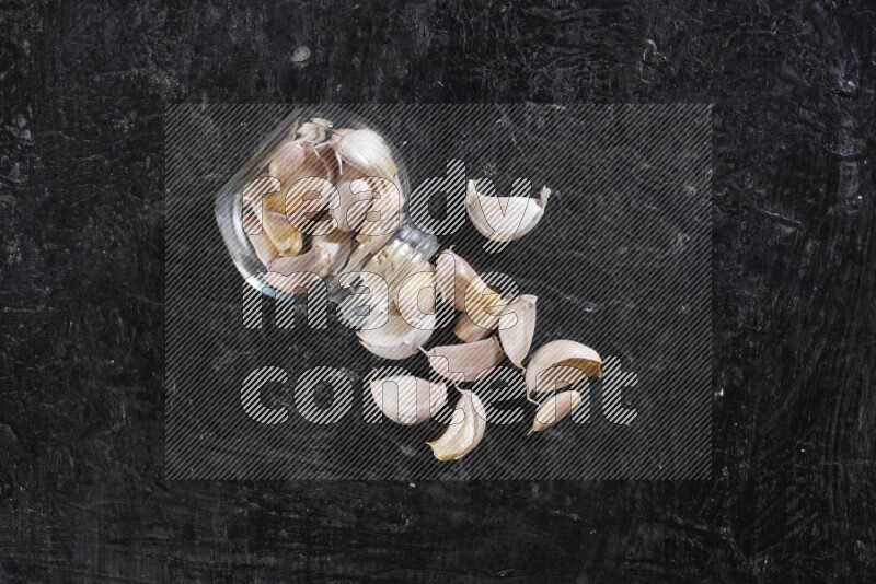 A glass spice jar full of garlic cloves flipped over with the cloves spilling out on a textured black flooring