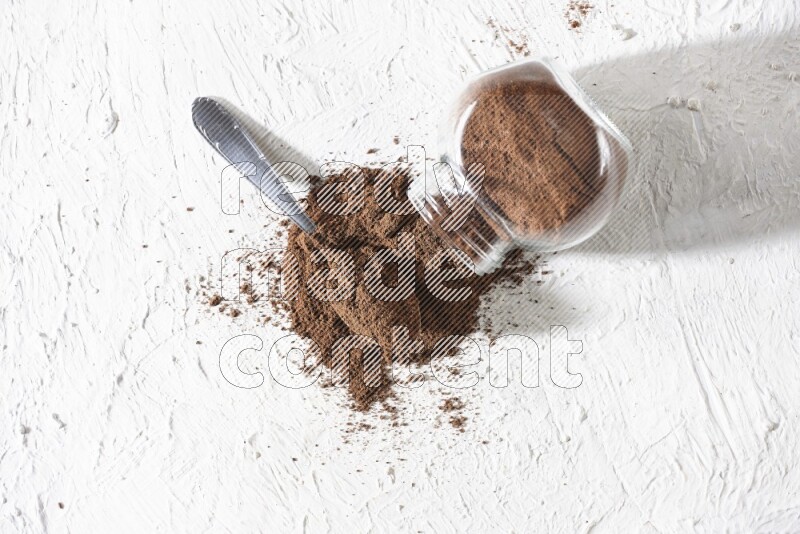 A flipped glass spice jar and a metal spoon full of cloves powder and powder came out of the jar on textured white flooring