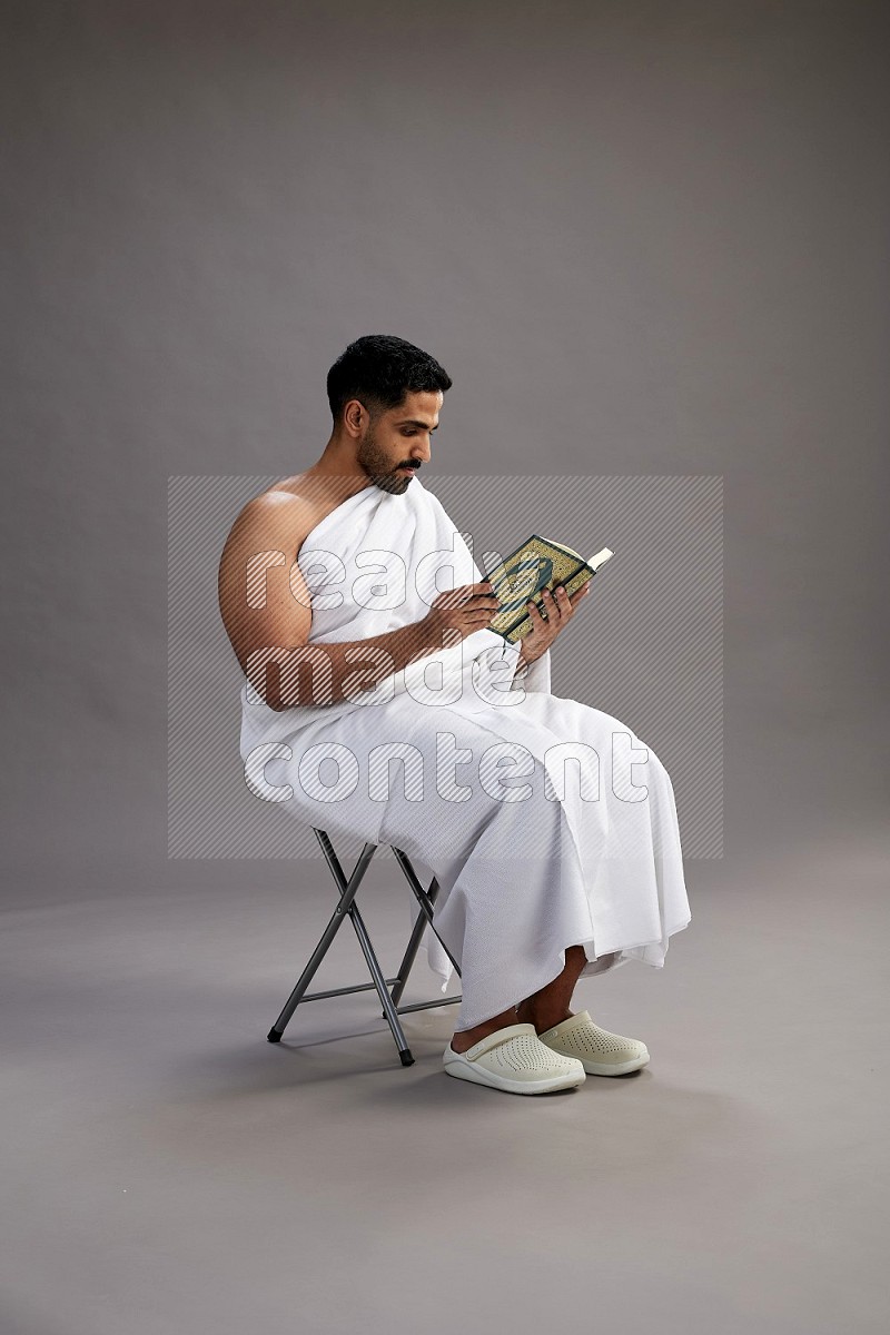 A man wearing Ehram sitting on chair reading quran on gray background