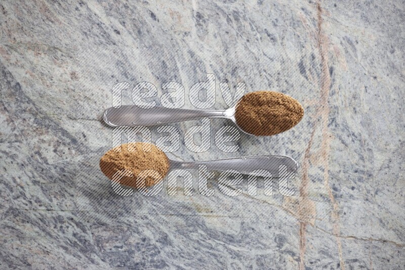 Two metal spoons full of cinnamon powder on grey marble background