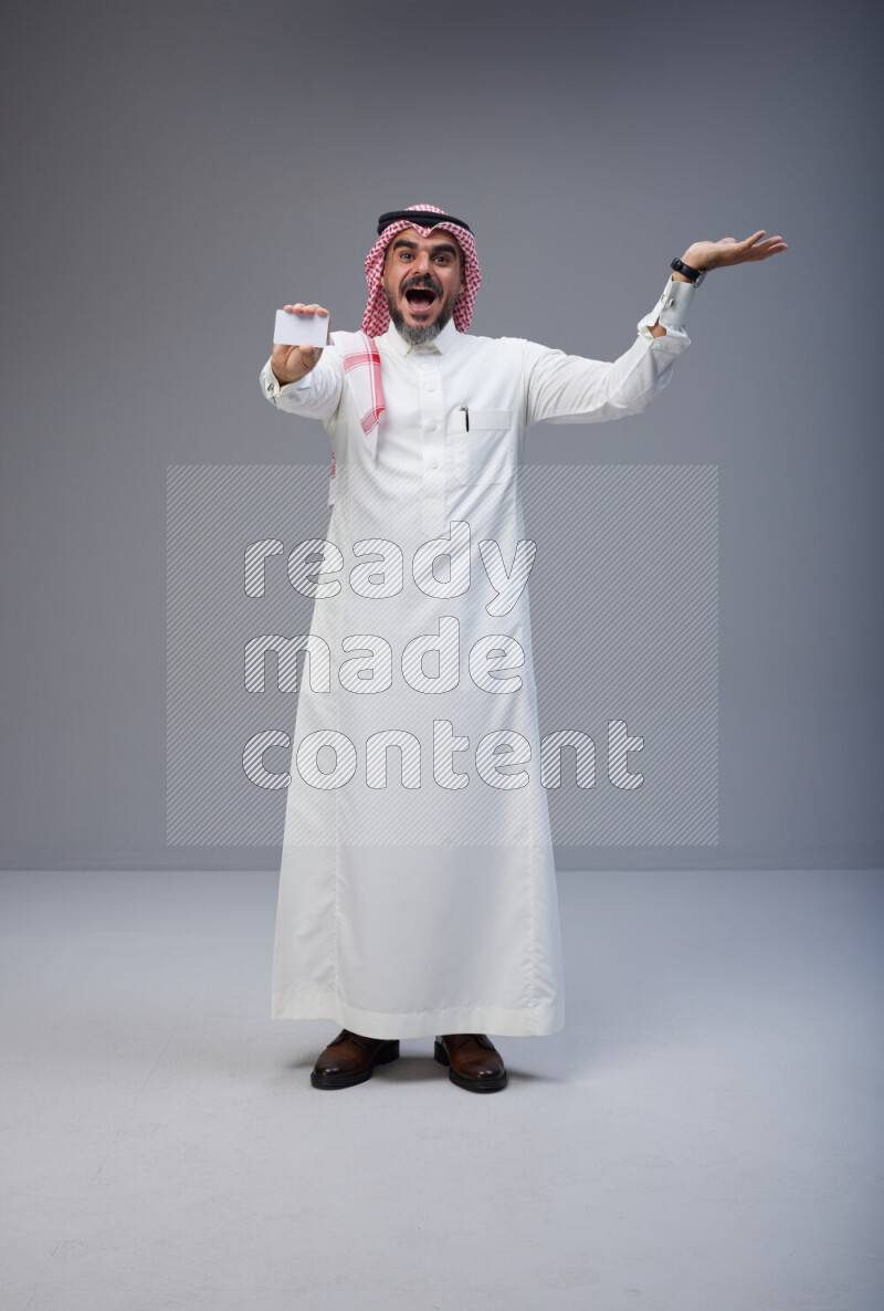 Saudi man Wearing Thob and red Shomag standing holding ATM card on Gray background