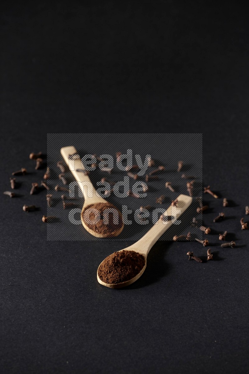 2 wooden spoons full of cloves powder with spreaded whole cloves on a black flooring