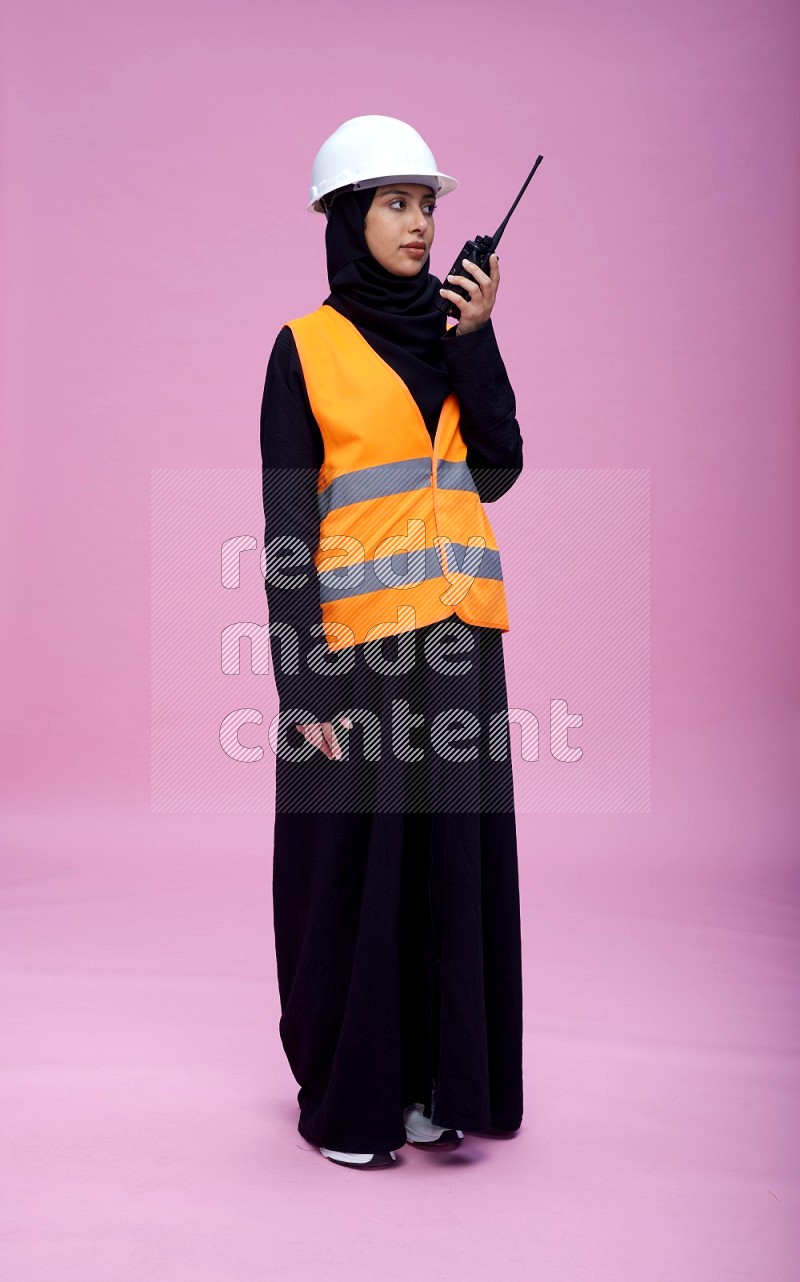 Saudi woman wearing Abaya with engineer vest and helmet standing holding walkie-talkie on pink background
