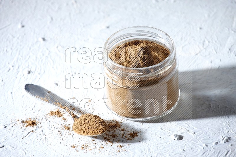 A glass jar and a metal spoon full of allspice powder on a textured white flooring