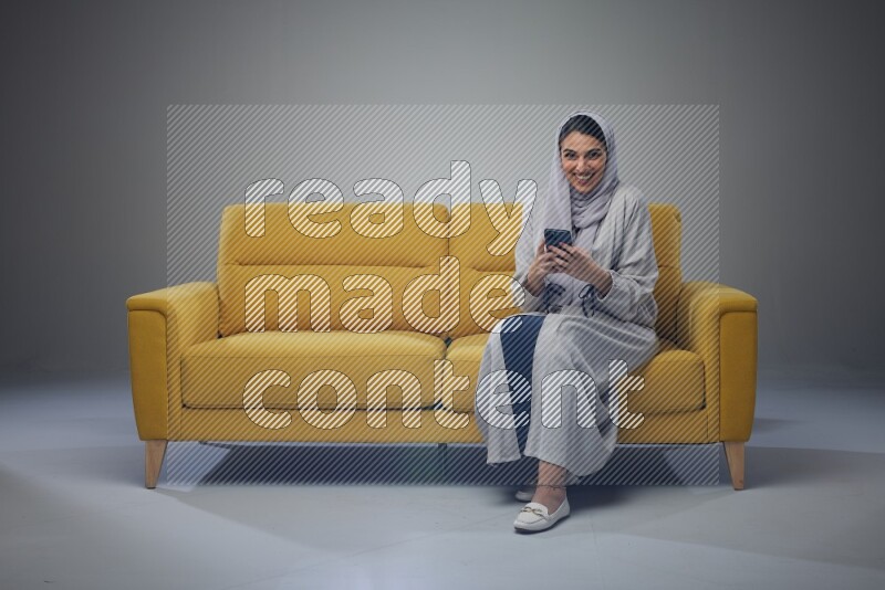 A Saudi woman wearing a light gray Abaya and white head scarf sitting on a yellow sofa and using her phone eye level on a grey background