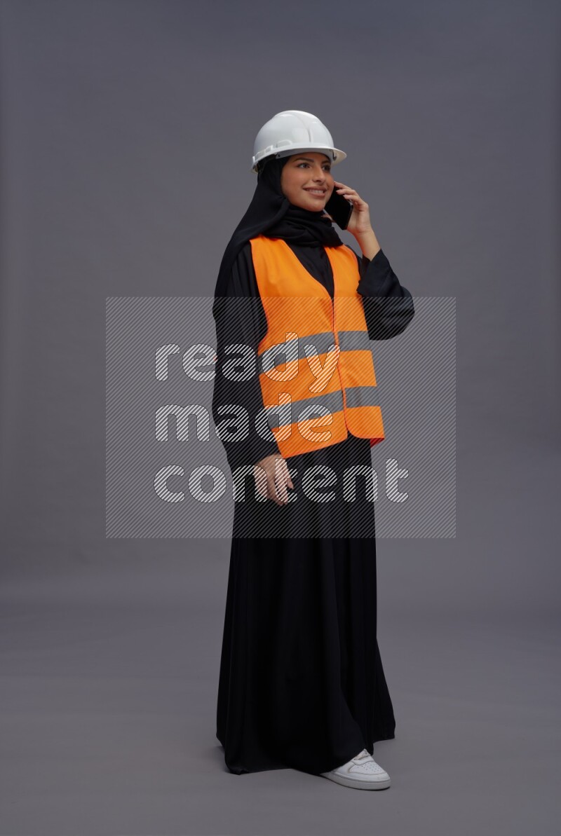 Saudi woman wearing Abaya with engineer vest standing talking on phone on gray background