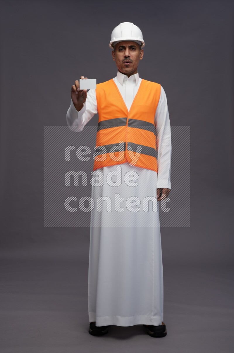 Saudi man wearing thob with engineer vest standing holding ATM card on gray background