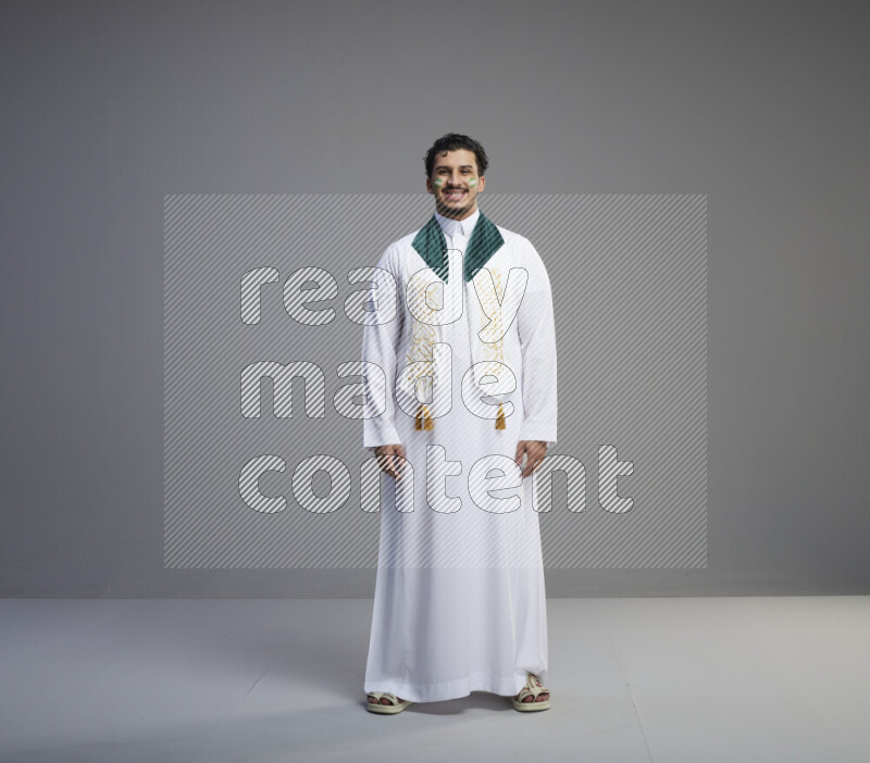 A Saudi man standing wearing thob and Saudi flag scarf with face painting on gray background