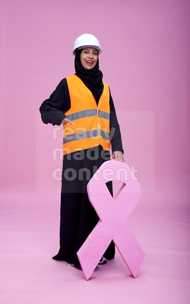 Saudi woman wearing Abaya with engineer vest and helmet standing awareness ribbon on pink background