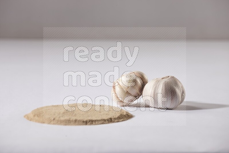 Garlic powder in a circle shape and beside it 2 garlic bulbs on a white flooring