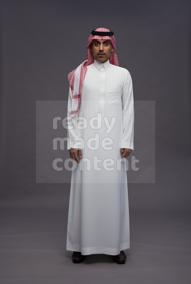 Saudi man wearing thob and shomag standing interacting with the camera on gray background