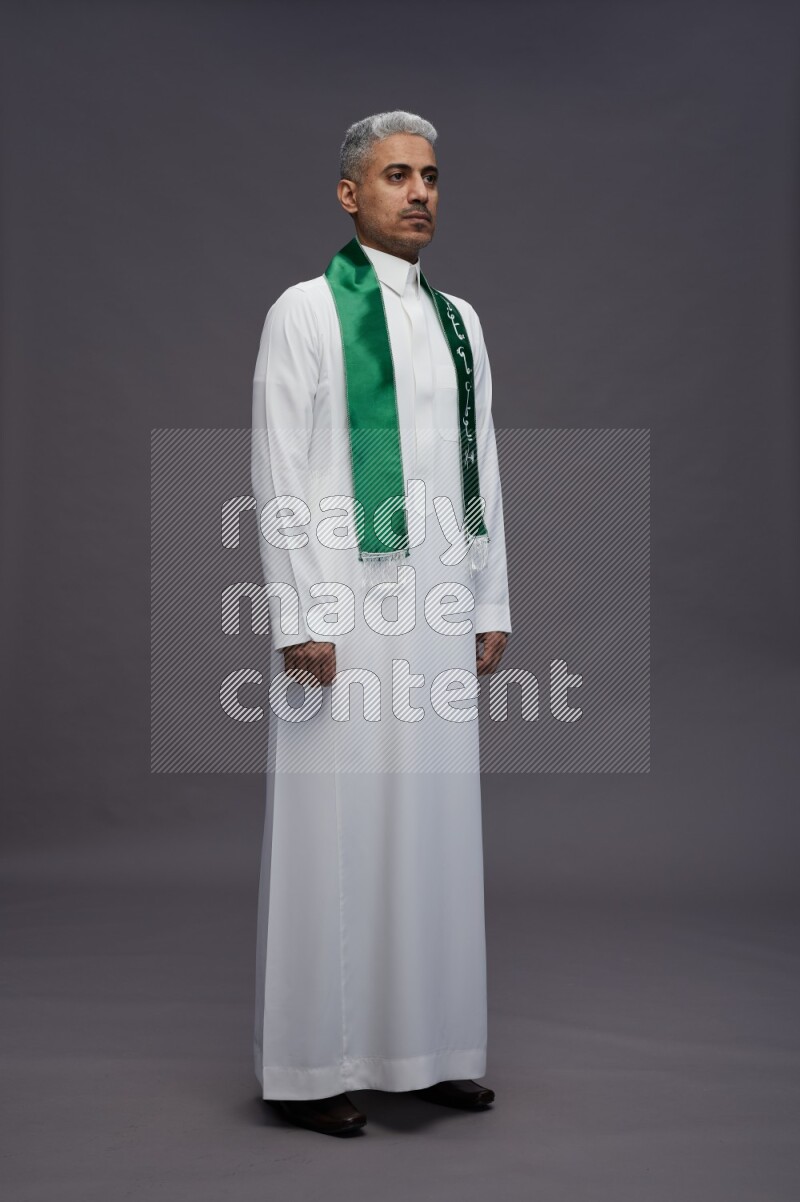 Saudi man wearing thob standing holding Saudi flag on gray background