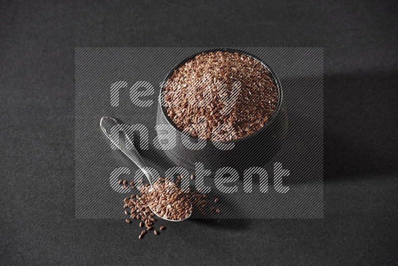 A black pottery bowl full of flaxseeds and a metal spoon full of the seeds on a black flooring