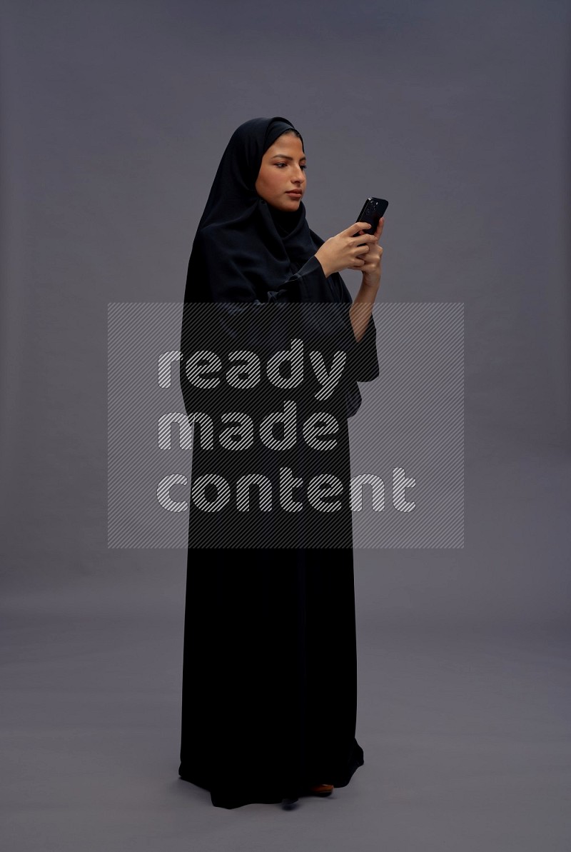 Saudi woman wearing Abaya standing texting on phone on gray background