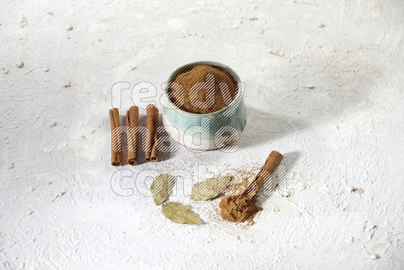 Cinnamon powder in a ceramic bowl with cinnamon sticks and laurel leaves on white background