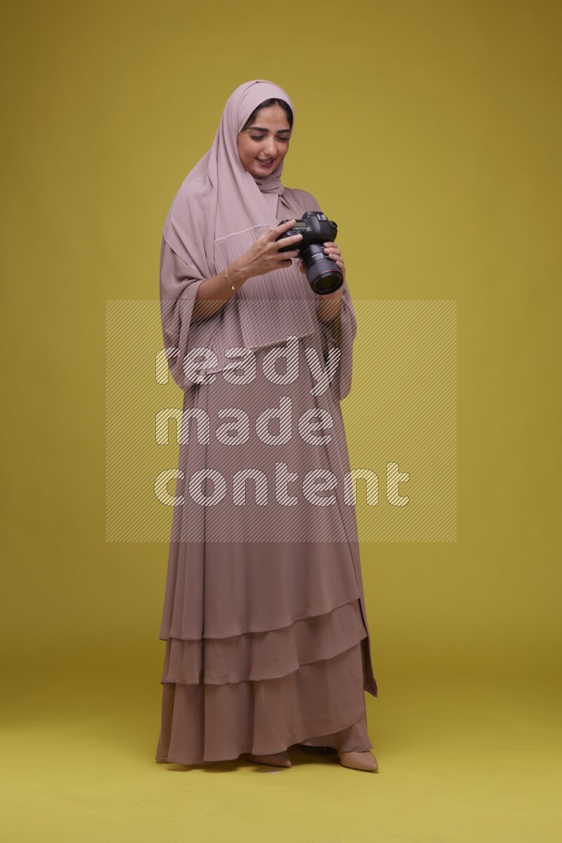 A woman Holding a Camera on a Yellow Background wearing Brown Abaya with Hijab