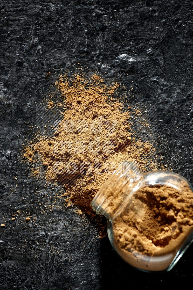 A flipped glass spice jar full of allspice powder and powder spilled out of it on a textured black flooring