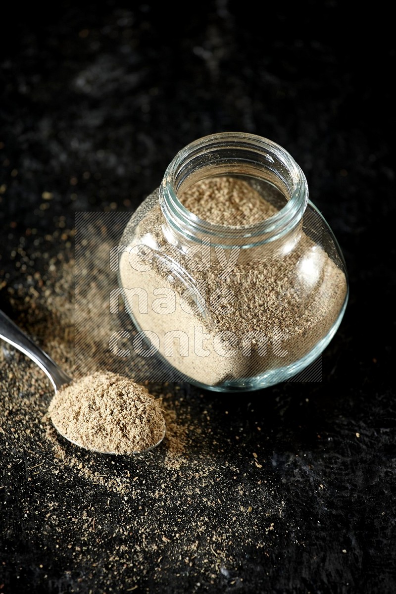 A glass spice jar and metal spoon full of cardamom powder on textured black flooring