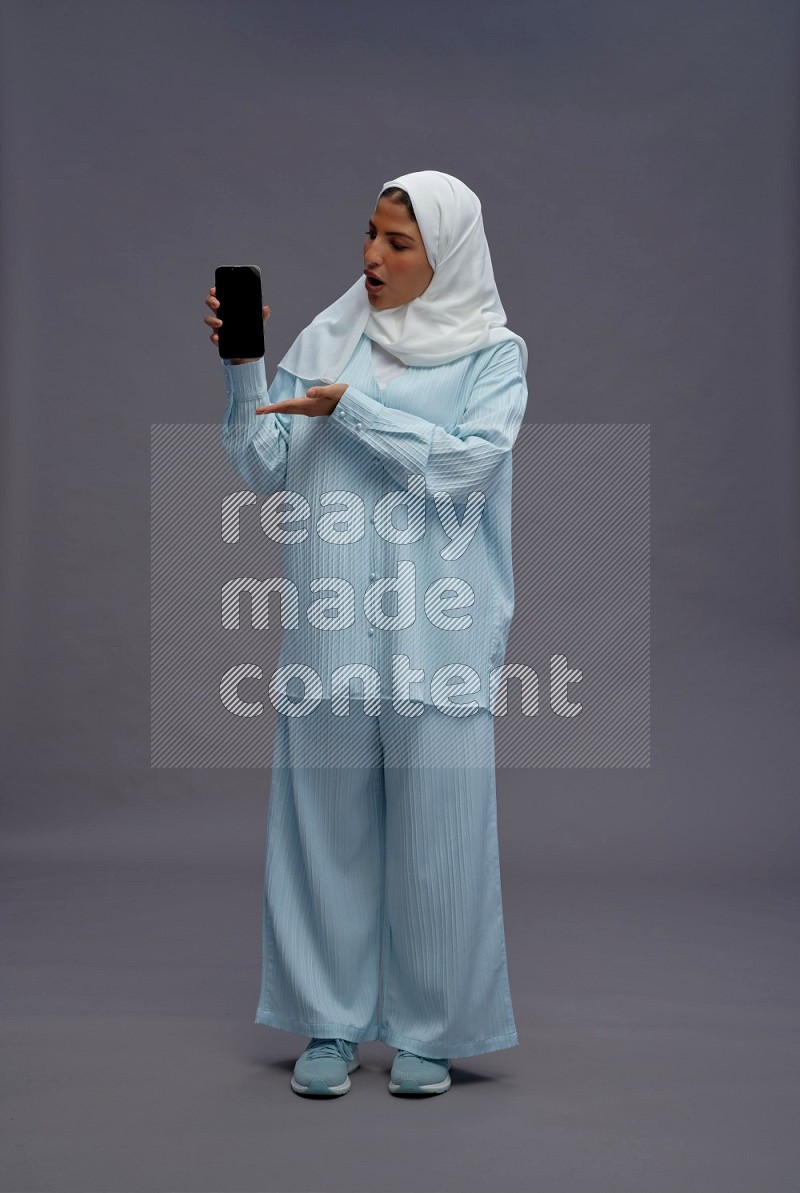 Saudi woman wearing hijab clothes standing showing phone to camera on gray background