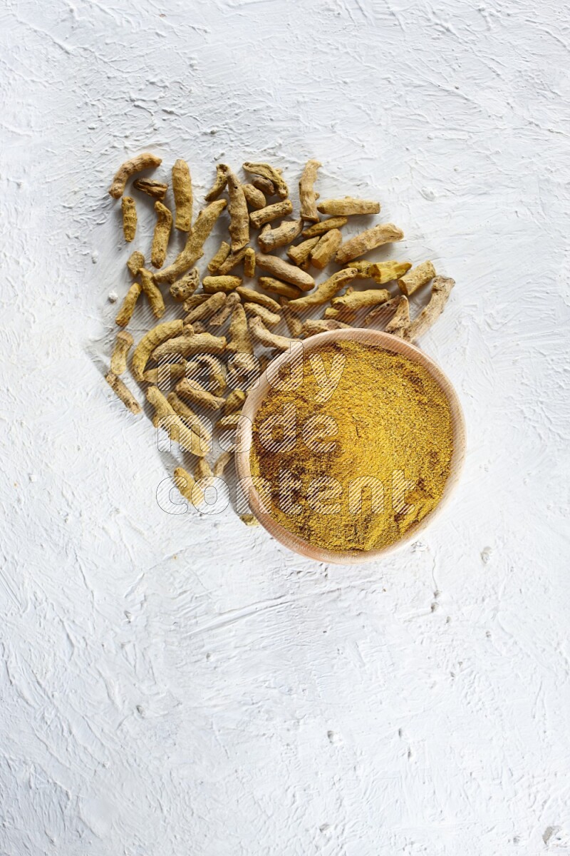 A wooden bowl full of turmeric powder and dried whole fingers beside it on a textured white flooring