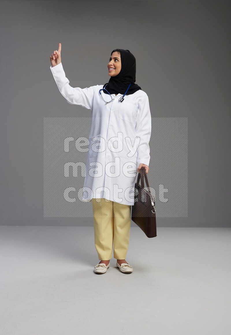 Saudi woman wearing lab coat with stethoscope standing holding bag on Gray background