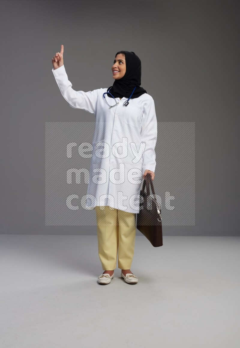 Saudi woman wearing lab coat with stethoscope standing holding bag on Gray background