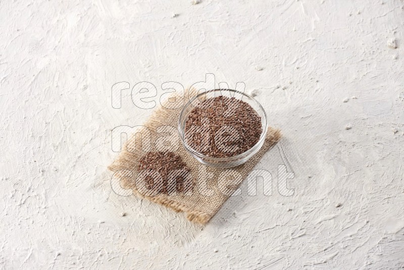 A glass bowl full of flax seeds and a bunch of seeds on burlap fabric on a textured white flooring