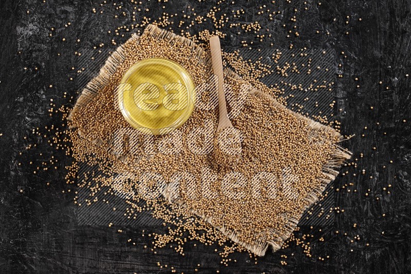 A glass jar full of mustard paste set on a burlap piece with a wooden spoon full of mustard seeds on a textured black flooring