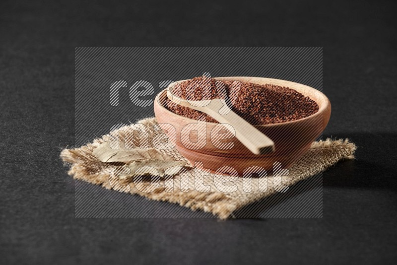 A wooden bowl full of garden cress seeds with wooden spoon full of the seeds on it on burlap fabric on a black flooring