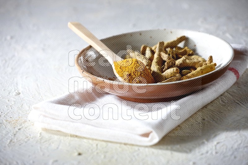 A plate filled with dried turmeric whole fingers and a wooden spoon full of turmeric powder on a textured white flooring